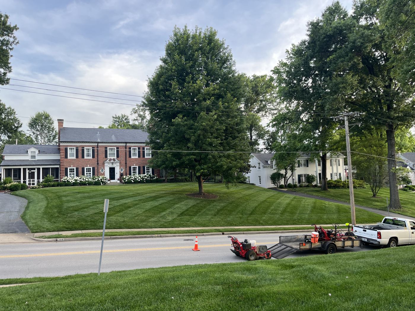 A red brick colonial with black shutters and white hydrangeas above a freshly striped front lawn. The Rolling Groves pickup, trailer, and two red mowers are parked at the curb.