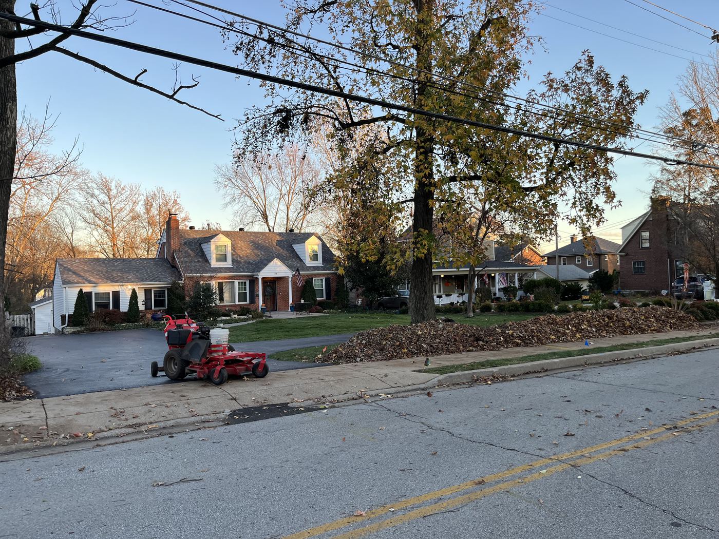 A brick Cape Cod house at golden hour. Fall leaves are raked into a long pile along the curb, and a red mower with a collector bag sits on the driveway.