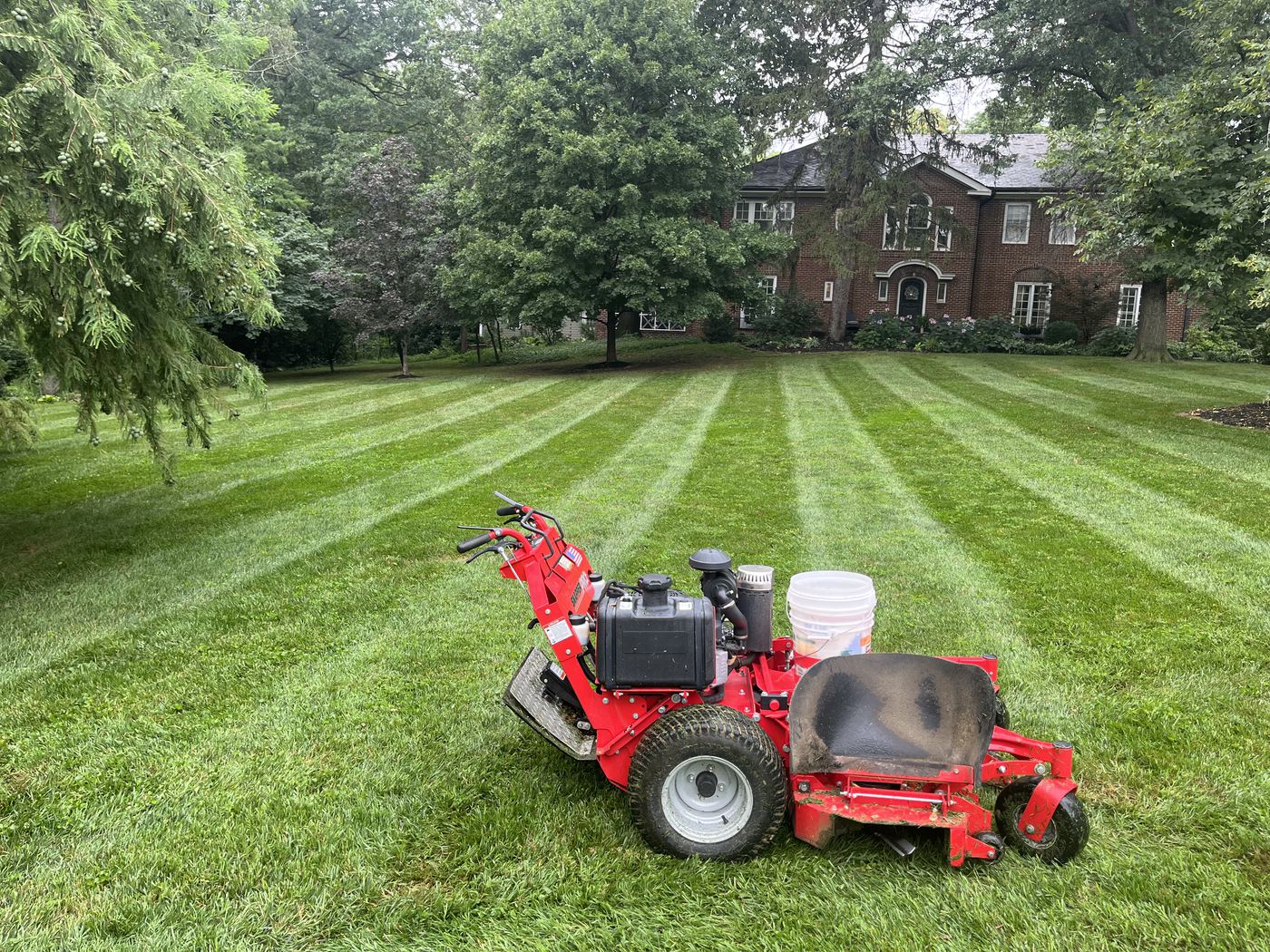 A red walk-behind mower with a white bucket parked on deep, crisp diagonal stripes, with a large brick house and mature trees behind.